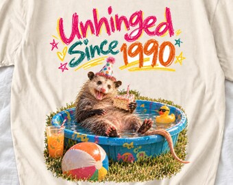 May include: A cream-colored t-shirt featuring a graphic of an opossum wearing a party hat in a blue kiddie pool. The opossum holds a slice of cake. Text reads "Unhinged Since 1990". A beach ball, a rubber duck, and a glass of orange juice are visible.