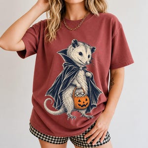 Etched pen illustration of an opossum in a vampire cape holding a candy pumpkin, bone white with orange accent