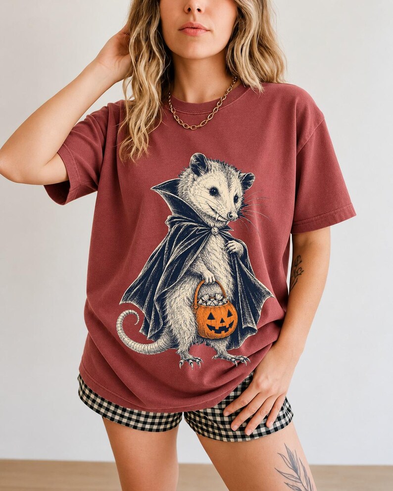 Etched pen illustration of an opossum in a vampire cape holding a candy pumpkin, bone white with orange accent