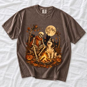 A skeleton in a scarf roasting marshmallows by a campfire beside a husky in an autumn forest under a full moon