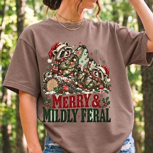 Two raccoons tangled in Christmas lights beside a log with the text Merry & Mildly Feral