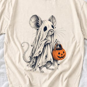 May include: A cream-colored t-shirt featuring a black and white ink-style illustration of a mouse wearing a tattered ghost costume. The mouse is holding a bright orange jack-o'-lantern candy bucket with a carved face.