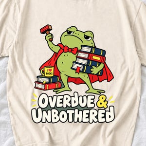 May include: A cream-colored t-shirt featuring a graphic of a green frog wearing a red cape, holding a gavel and a stack of books. A yellow mug labeled I heart books sits on a stack of books. The text reads OVERDUE & UNBOTHERED in bold letters.