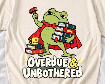 Puede incluir: Una camiseta color crema con el gráfico de una rana verde con capa roja, sosteniendo un martillo y libros. Una taza amarilla con la etiqueta I heart books descansa sobre una pila de libros. El texto dice OVERDUE & UNBOTHERED en letras negritas.