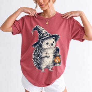 Etched pen illustration of a hedgehog in a witch hat holding a lantern, bone white with orange accent