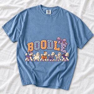 Six poodles in Halloween costumes walking under the arched word BOODLE, lavender and orange polka-dot varsity style