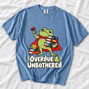 Grumpy cartoon frog librarian holding a stamp and a stack of books with the text Overdue & Unbothered