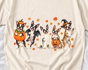 May include: A cream-colored t-shirt featuring a graphic print of five Boston Terrier dogs dressed in Halloween costumes, including a pumpkin, bat wings, a ghost sheet, and a witch hat, surrounded by scattered orange autumn leaves.