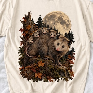 May include: A cream-colored t-shirt featuring a graphic print of a large opossum carrying three smaller opossums on its back. The scene includes a forest background with a large full moon, autumn leaves, and a mossy tree trunk.