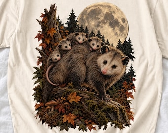 May include: A cream-colored t-shirt featuring a graphic print of a large opossum carrying three smaller opossums on its back. The scene includes a forest background with a large full moon, autumn leaves, and a mossy tree trunk.