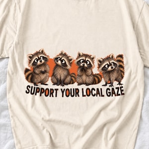 May include: A cream-colored t-shirt featuring a graphic of four illustrated raccoons standing in a row against an orange background. Below the raccoons, the text "SUPPORT YOUR LOCAL GAZE" is printed in bold, dark brown capital letters.
