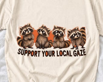 May include: A cream-colored t-shirt featuring a graphic of four illustrated raccoons standing in a row against an orange background. Below the raccoons, the text "SUPPORT YOUR LOCAL GAZE" is printed in bold, dark brown capital letters.