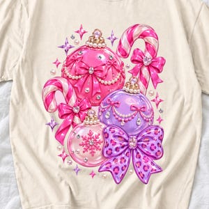 May include: A cream-colored t-shirt featuring a graphic print of three Christmas ornaments in pink and purple, two candy canes, and bows. The ornaments are decorated with pearls, rhinestones, and a snowflake pattern, while a large purple bow features a leopard print design.