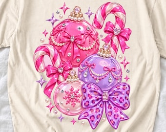 May include: A cream-colored t-shirt featuring a graphic print of three Christmas ornaments in pink and purple, two candy canes, and bows. The ornaments are decorated with pearls, rhinestones, and a snowflake pattern, while a large purple bow features a leopard print design.