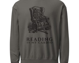 Op de afbeelding: Een antracietgrijs sweatshirt met ronde hals en een zwarte afbeelding van een skelet dat in een fauteuil zit en een boek leest, met een paar sneakers op de grond. De tekst onder de afbeelding luidt "READING IS MY CARDIO" in een schreeflettertype.
