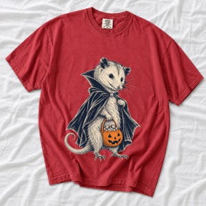Etched pen illustration of an opossum in a vampire cape holding a candy pumpkin, bone white with orange accent