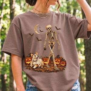 A skeleton walking a corgi in a ghost costume past carved pumpkins under a crescent moon