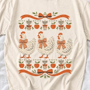 Three fluffy hens with gingham bows between apple and wheat borders, retro folk art fall t-shirt design