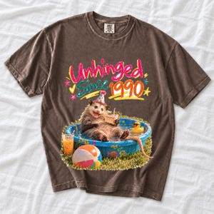 Opossum in party hat in a kiddie pool with cake, 90s film photo style, text "Unhinged Since 1990"