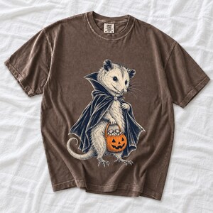 Etched pen illustration of an opossum in a vampire cape holding a candy pumpkin, bone white with orange accent