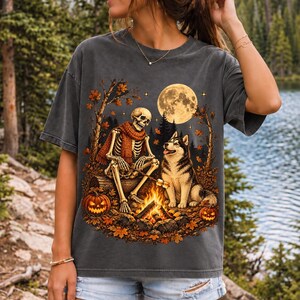 A skeleton in a scarf roasting marshmallows by a campfire beside a husky in an autumn forest under a full moon