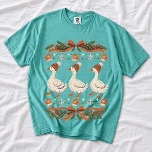 Three geese in pompom knit beanies with mittens, snowflakes and pine borders, retro winter t-shirt graphic