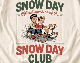 May include: A cream-colored t-shirt featuring a graphic of two children and a dog on a wooden sled. The text reads SNOW DAY, Official members of the, and SNOW DAY CLUB in green and red lettering. The illustration uses a vintage color palette.