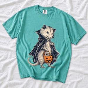 Etched pen illustration of an opossum in a vampire cape holding a candy pumpkin, bone white with orange accent