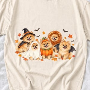 May include: A cream-colored t-shirt featuring a graphic of five Pomeranian dogs dressed in Halloween costumes, including a witch, a ghost, a pumpkin, a lion, and a bat, surrounded by scattered orange autumn leaves and small black bats.