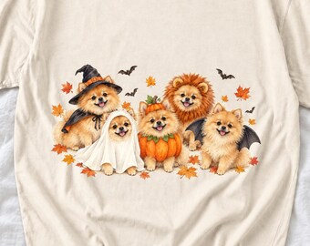 May include: A cream-colored t-shirt featuring a graphic of five Pomeranian dogs dressed in Halloween costumes, including a witch, a ghost, a pumpkin, a lion, and a bat, surrounded by scattered orange autumn leaves and small black bats.