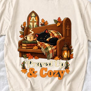 Black cat curled on a church pew with blanket and cocoa among autumn leaves, text "Prayed Up & Cozy", retro illustration
