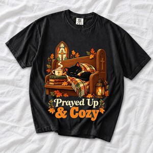 Black cat curled on a church pew with blanket and cocoa among autumn leaves, text "Prayed Up & Cozy", retro illustration