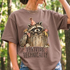 Raccoon in a paper party hat peeking out of a trash can with the text "Thriving, Technically", sepia watercolor sketch