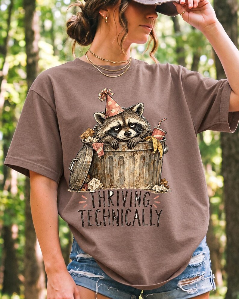 Raccoon in a paper party hat peeking out of a trash can with the text "Thriving, Technically", sepia watercolor sketch