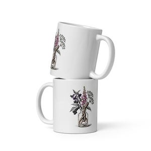 Op de afbeelding: Twee witte keramische mokken verticaal gestapeld. Elke mok is voorzien van een botanische illustratie van een glazen fles met daarin een kleine schedel en een boeket van paarse vingerhoedskruidbloemen, donkere bessen en groen blad.