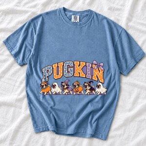 Six pugs in pumpkin, ghost and witch costumes under the arched word PUGKIN, lavender and orange polka-dot varsity style