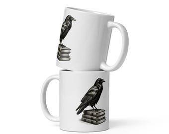 May include: Two white ceramic mugs stacked vertically, each featuring a detailed black and white illustration of a raven perched atop a small stack of three books. The mugs have a smooth finish, a curved handle on the side, and a cylindrical shape.