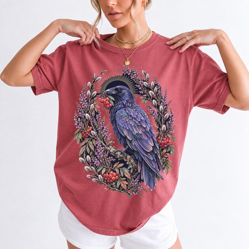 Raven holding a rowan berry inside an oval wreath of heather and catkins, painted cottagecore bird illustration
