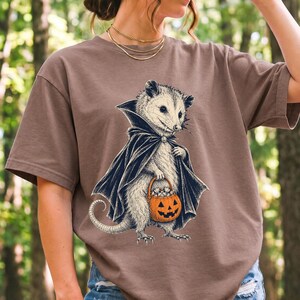 Etched pen illustration of an opossum in a vampire cape holding a candy pumpkin, bone white with orange accent