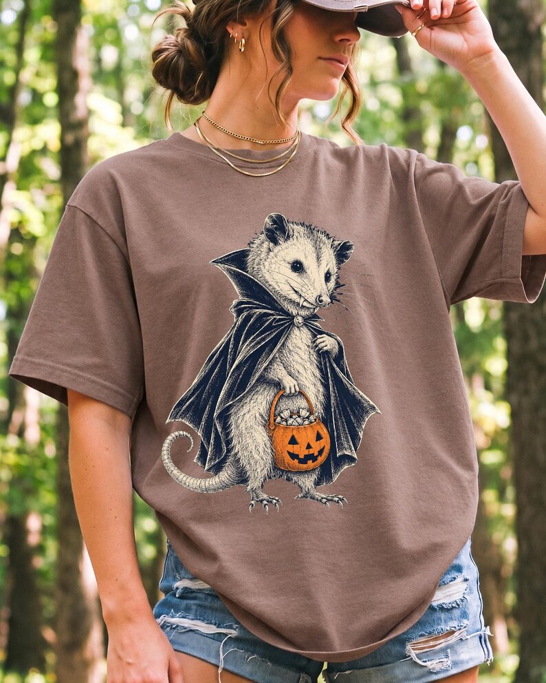 Etched pen illustration of an opossum in a vampire cape holding a candy pumpkin, bone white with orange accent