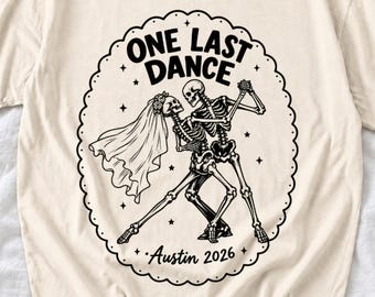 May include: A cream-colored t-shirt featuring a black graphic of two skeletons dancing. The design is enclosed in a scalloped oval border with the text "ONE LAST DANCE" at the top and "Austin 2026" at the bottom, accented by small stars.