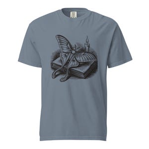 May include: A slate blue short-sleeve t-shirt featuring a black and white graphic print on the chest. The illustration depicts a large moth resting on top of a closed book, positioned next to a single lit candle with a visible flame and wax drips.