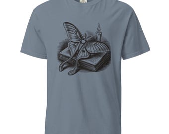 Puede incluir: Una camiseta de manga corta color azul pizarra con un estampado gráfico en blanco y negro en el pecho. La ilustración muestra una polilla grande sobre un libro cerrado, junto a una vela encendida con una llama visible y gotas de cera.