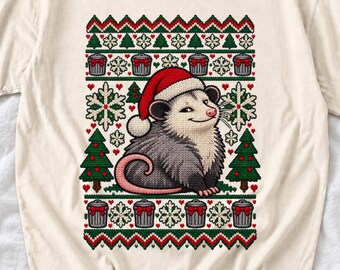 May include: A cream-coloured t-shirt featuring a graphic of an opossum wearing a red and white Santa hat. The design includes a festive pattern of green pine trees, white snowflakes, red hearts, and grey trash cans with red bows.