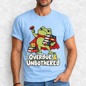 Grumpy cartoon frog librarian holding a stamp and a stack of books with the text Overdue & Unbothered