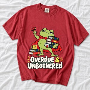 Grumpy cartoon frog librarian holding a stamp and a stack of books with the text Overdue & Unbothered