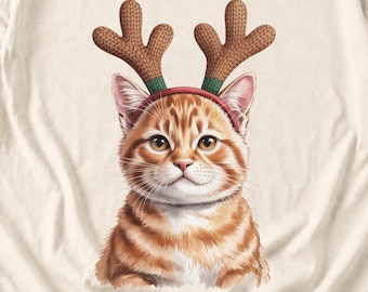 May include: A cream-colored t-shirt featuring a graphic illustration of an orange tabby cat wearing a red headband with brown, textured, knit-style reindeer antlers. The cat has large, expressive eyes and detailed whiskers.