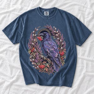 Raven holding a rowan berry inside an oval wreath of heather and catkins, painted cottagecore bird illustration