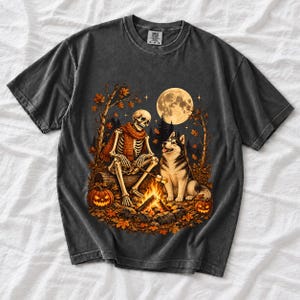 A skeleton in a scarf roasting marshmallows by a campfire beside a husky in an autumn forest under a full moon