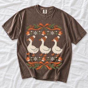 Three geese in pompom knit beanies with mittens, snowflakes and pine borders, retro winter t-shirt graphic